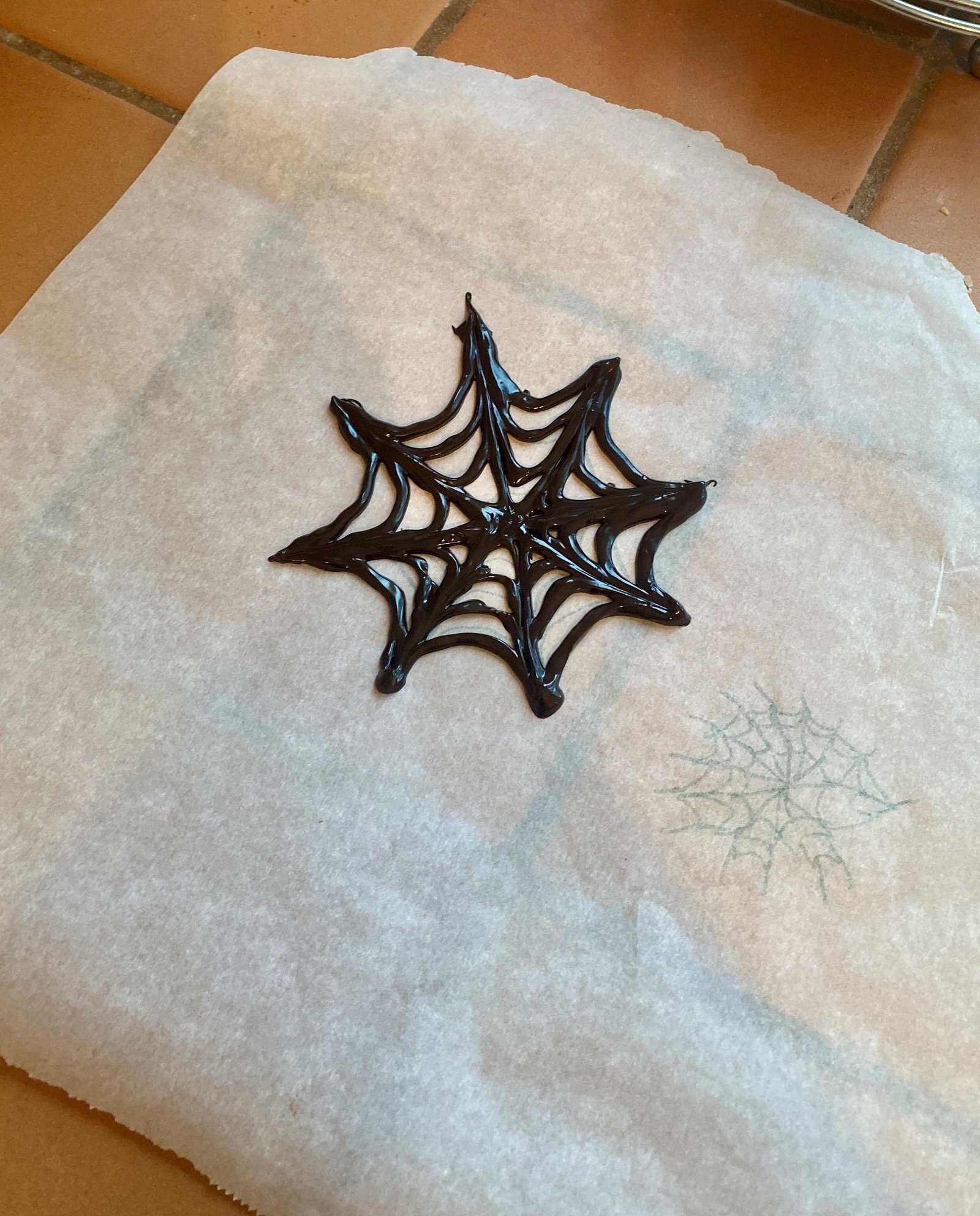 A glossy chocolate web piped on parchment, with a pencil spiderweb guide visible nearby through the paper.